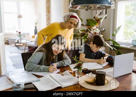 Seniorin hilft Enkelkindern bei Hausaufgaben, während sie zu Hause sitzen Stockfoto