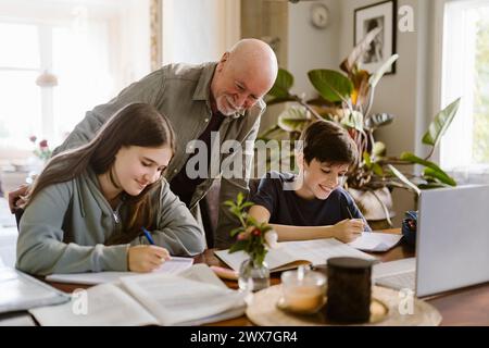 Lächelnder Großvater, der Enkel ansieht, wie sie Hausaufgaben machen, während er zu Hause am Tisch sitzt Stockfoto