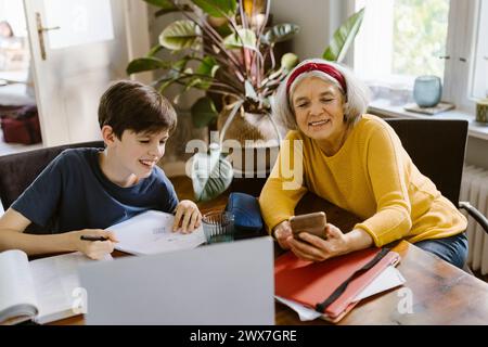 Lächelnde Seniorin, die ihr Smartphone benutzt, während sie mit Enkel zu Hause Hausaufgaben macht Stockfoto