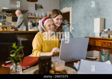 Lächelnde Enkelin, die Großmutter mit Laptop zu Hause umschließt Stockfoto