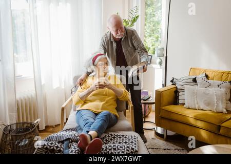 Senior-Mann, der der Frau, die auf einem Stuhl sitzt und zu Hause den Becher hält, Buch zeigt Stockfoto