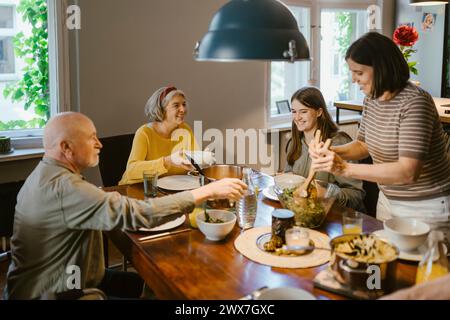 Lächelnde Frau, die der Familie Salat serviert, während sie zu Hause am Esstisch zu Abend essen Stockfoto