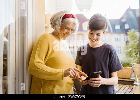 Lächelnde ältere Frau, die das Smartphone benutzt, das von Enkel gehalten wird, der auf dem Balkon steht Stockfoto