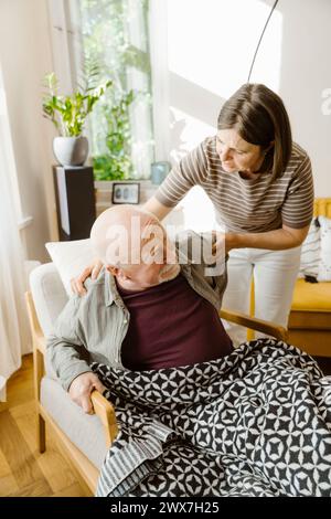 Reife Frau kümmert sich um einen älteren Mann, der zu Hause auf einem Stuhl sitzt Stockfoto