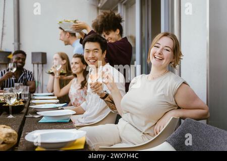 Porträt einer glücklichen jungen Frau, die Weinglas hält, während sie mit einer Gruppe von Freunden auf der Abendparty auf dem Balkon sitzt Stockfoto
