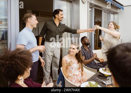 Lächelnde Frau, die Weingläser an einen männlichen Freund übergibt, auf einer Dinnerparty auf dem Balkon Stockfoto