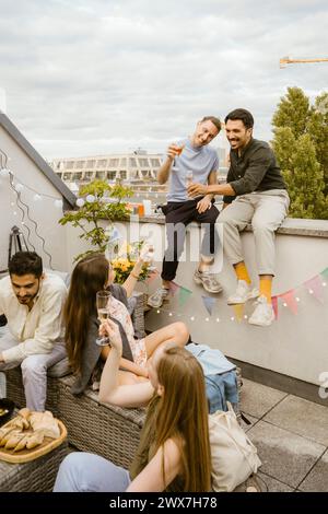 Lächelnde Männer, die mit Freunden Getränke trinken, während sie auf dem Balkon sitzen Stockfoto