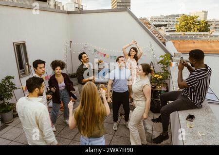 Junger Mann, der männliche und weibliche Freunde fotografiert, die auf der Dachterrasse auf einer Party tanzen Stockfoto