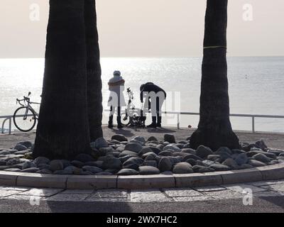 Ein Paar mit ihren Fahrrädern hat eine Silhouette in der Wintersonne vor dem glitzernden Mittelmeer auf der Promenade des Anglais in Nizza. Stockfoto