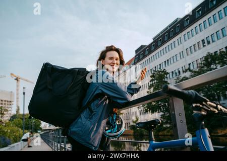 Seitliches Porträt einer lächelnden weiblichen Lieferperson, die in der Stadt ihr Smartphone hält, während sie sich auf das Geländer stützt Stockfoto