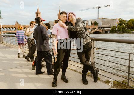 Schwuler Mann lehnt sich an einen nicht-binären Freund, der neben einem Geländer an der Promenade in der Stadt steht Stockfoto