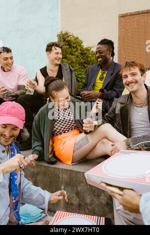 Glückliche, nicht-binäre Freunde, die auf der Bank mit Essen und Getränken feiern Stockfoto