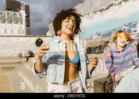 Unbeschwert lächelnde, nicht-binäre Person, die mit Freunden tanzt, die an sonnigen Tagen auf Stufen in der Stadt sitzen Stockfoto