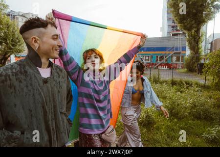 Frau, die LGBT-Flagge hält, während sie mit nicht-binären Freunden im Park läuft Stockfoto