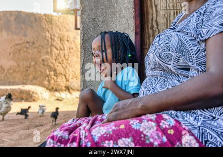 Verschlafenes, müdes afrikanisches Kind mit Zöpfen, die zusammen mit ihrer Oma auf der Veranda sitzen, Hühner rennen, afrikanisches Dorfleben Stockfoto