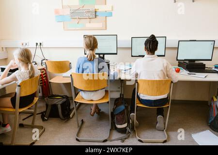 Rückansicht multirassischer Schülerinnen, die Computer benutzen, während sie in der Schule auf Stühlen sitzen Stockfoto