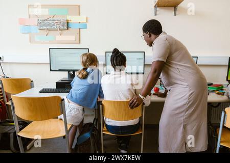 Seitenansicht einer Lehrerin, die Mädchen mit Computer erklärt, während sie in der Schule sitzen Stockfoto