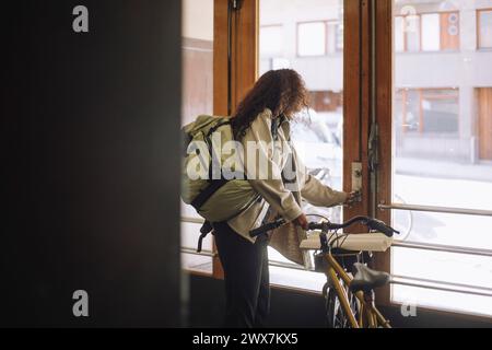 Seitenansicht der freiberuflich arbeitenden Frau, die beim Verlassen des Gebäudes mit dem Fahrrad die Tür öffnet Stockfoto