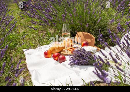 Ein Sommer-Picknick in einem Lavendelfeld mit Croissants, Pfirsichen, Salami, Käse und einer Flasche Wein und Gläsern. Violette Blumen auf dem Hintergrund. Stockfoto