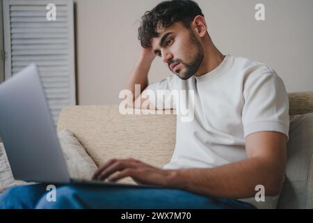 Porträt eines attraktiven jungen bärtigen Mannes, der legere Kleidung trägt und im Wohnzimmer auf dem Sofa sitzt und mit einem Laptop-Computer in sozialen Netzwerken chatten kann Stockfoto