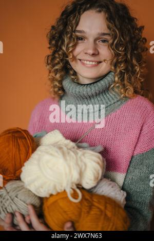 Lockenhaarige Frau Porträt mit Strickfäden, isoliert auf orangem Hintergrund. Handgefertigtes Hobby-Stay-Home-Konzept. Craft und DIY-Konzept. Stockfoto