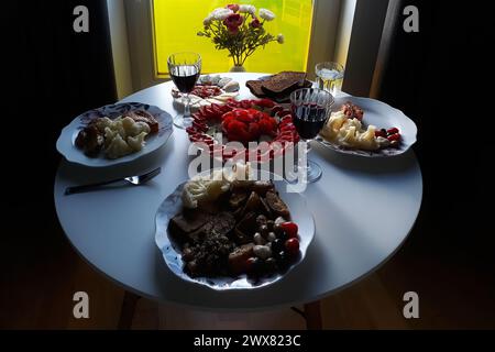 Ein runder Tisch mit Gläsern Wein und verschiedenen Speisen und Snacks in gemütlicher Halbdunkel, beleuchtet durch Licht aus dem Fenster. Stockfoto