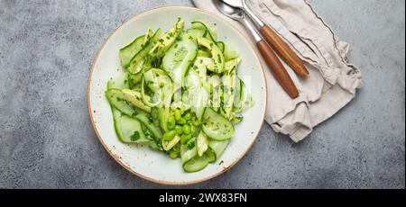 Gesunde vegane grüne Avocado Salatschüssel mit Gurkenscheiben, Edamam-Bohnen, Olivenöl und Kräutern auf Keramikplatte, Blick von oben, grauer Stein rustikaler Tisch Stockfoto