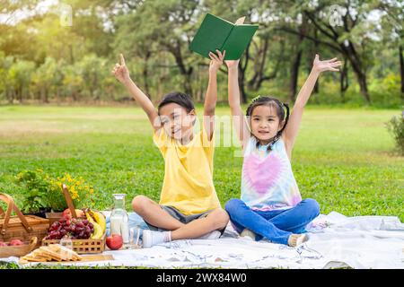 Zwei Kinder sitzen im Gras, lesen und heben die Hände Stockfoto