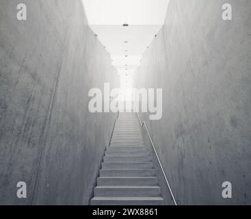 Schmale Betonstufen, die zum Licht führen. Leere Treppe mit Geländern auf der rechten Seite, Betonwände und beleuchtete satinierte Glasdecke. Stockfoto