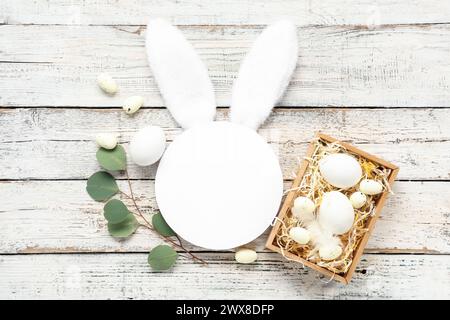 Leere Karte mit Hasenohren, Korb und Ostereiern auf weißem hölzernem Hintergrund Stockfoto