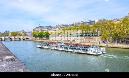 Ein mit Touristen gefülltes Flussboot schwimmt entlang der seine in Paris, mit Stadtgebäuden und einer Brücke im Hintergrund. Frankreich Stockfoto