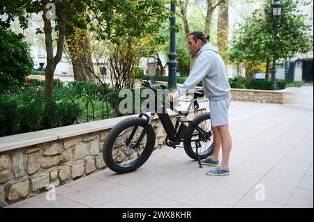 Ein junger Mann, der ein Elektrofahrrad für die Fahrt in der Stadt mietet. Elektrofahrrad - Öko-öffentliche Verkehrsmittel. Stadtservice für Fahrradgemeinschaften. Umgebung Stockfoto