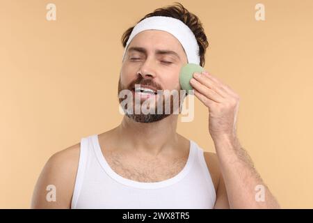 Mann mit Stirnband, der sein Gesicht mit einem Schwamm auf beigem Hintergrund wäscht Stockfoto
