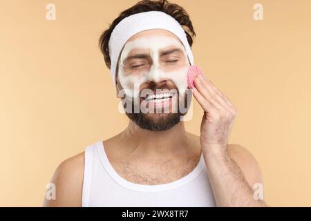 Mann mit Stirnband, der sein Gesicht mit einem Schwamm auf beigem Hintergrund wäscht Stockfoto