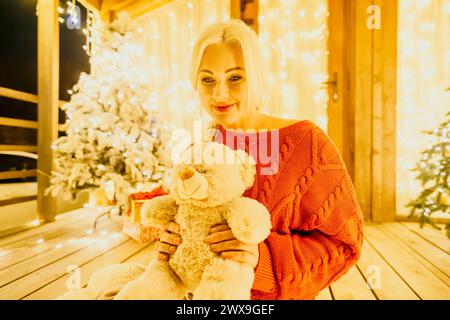 Eine glückliche lächelnde Frau in einem roten Pullover sitzt auf einer festlichen Veranda und hält einen Teddybären neben einem schneebedeckten Weihnachtsbaum mit hellen weißen Lichtern Stockfoto