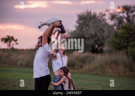 Vielfalt glückliche Familie spielt bei Sonnenuntergang auf dem Feld. Junger Vater hebt das Kleinkind hoch in die Luft, während die ältere Geschwistertochter und Mutter mit Feinkost beobachten Stockfoto