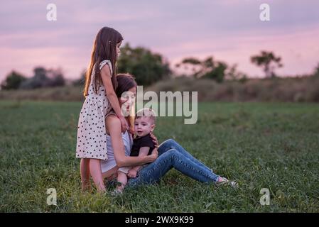 Mutter umarmt Sohn und Tochter, sitzt auf Gras, während die Dämmerung sich einsetzt. Moment der stillen Zuneigung, Verbindung. Ruhige Familienzeit in der Abenddämmerung. W Stockfoto