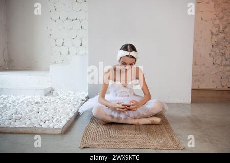 Junge Ballerina in weißem Tutu, mit Federdiadem, sitzt auf dem Boden im Loft-Innenraum, hält das Smartphone in den Händen, surft im Internet. Soziales Teenager Stockfoto