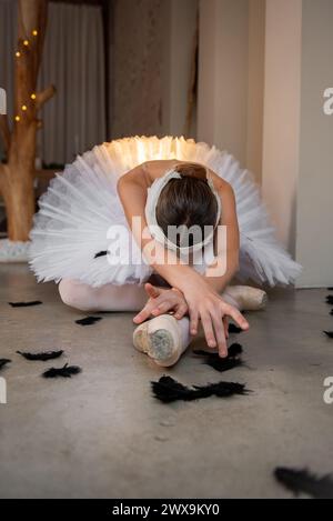 Die Ballerina zeigt den sterbenden Schwan im Swan Lake Ballet. Balletttänzerin in spitzenschuhen liegt auf dem Boden zwischen schwarz gefallenen Federn. Talentiertes Theater A Stockfoto