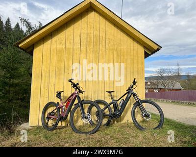 Zwei moderne E-Mountainbikes, die sich an ein kleines Holzhaus lehnen (keine Leute). Stockfoto