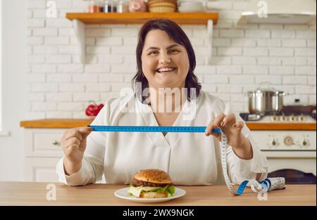 Eine fette, übergewichtige junge Frau, die in der Küche sitzt und ein Maßband mit einem Burger auf dem Tisch hält. Stockfoto