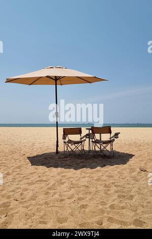 Rayong Thailand 13. März 2024, zwei Stühle sitzen unter einem bunten Sonnenschirm an einem ruhigen Sandstrand, laden zum Entspannen ein und genießen den malerischen Meerblick. Stockfoto