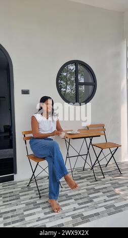 Eine Frau sitzt friedlich an einem Tisch vor einem charmanten Haus, umgeben von Natur, verloren in Gedanken. Stockfoto