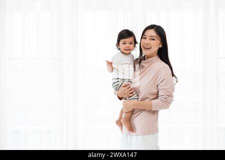 Glückliche Mutter, die Kleinkind auf weißem Fensterhintergrund hält Stockfoto