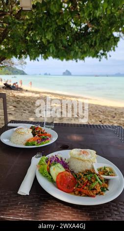 Zwei Teller mit köstlichem Essen stehen auf einem Tisch in der Nähe des Strandes, wobei die Wellen des Ozeans sanft unter dem blauen Himmel in den Hintergrund stürzen. Koh Kradan Thailand Stockfoto