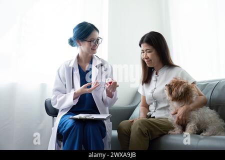 Ältere Frau mit Hund sitzt am Schreibtisch vor asiatischer Tierärztin in der Tierarztpraxis Stockfoto