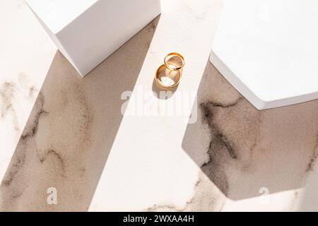Zwei klassische goldene Eheringe liegen auf einem beigefarbenen Marmorhintergrund zwischen zwei weißen geometrischen Podesten von Figuren und Sonnenstrahlen. Draufsicht Stockfoto