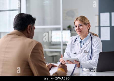 Porträt einer lächelnden Ärztin, die mit dem Mann aus dem Nahen Osten während der Sprechstunde in der medizinischen Klinik spricht Stockfoto