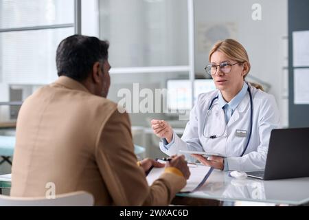 Porträt einer lächelnden Ärztin, die einen Mann aus dem Nahen Osten berät, der in der medizinischen Klinik über den Tisch sitzt Stockfoto