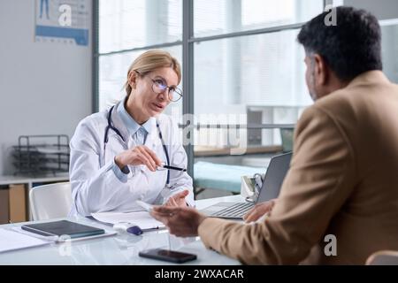 Porträt einer reiferen Ärztin mit Brille und Gespräch mit dem Patienten über den Tisch in der medizinischen Klinik Stockfoto